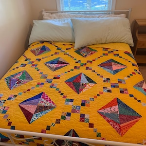 May include: A bed featuring a bright yellow quilt with a patchwork design of diamond-shaped blocks in various colorful patterns, connected by small multi-colored square segments. Two light blue pillows rest at the head of the white metal bed frame.