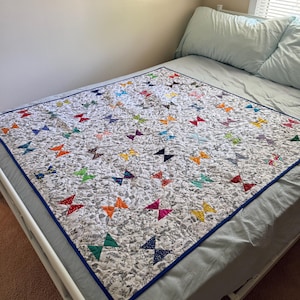 Può includere: Una trapunta rettangolare con sfondo bianco e un denso motivo floreale nero e grigio. La trapunta è decorata con numerose piccole forme a farfalla colorate in varie tonalità, tra cui rosso, arancione, giallo, verde, blu e viola.