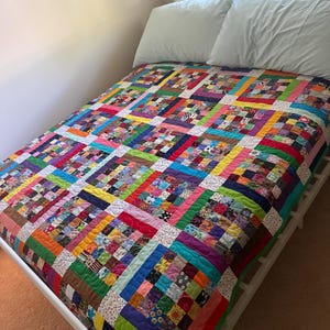 Könnte beinhalten: Eine Patchwork-Decke mit einem Raster aus bunten Stoffquadraten in verschiedenen Mustern, die durch einfarbige Stoffränder in Rot, Blau, Grün und Gelb getrennt sind, mit weiß gemusterten Zwischenstreifen zwischen den einzelnen Blöcken.
