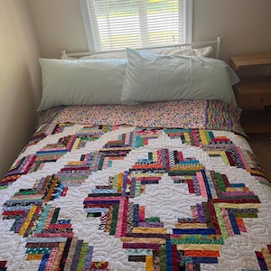 May include: A bed featuring a log cabin style quilt with a white background and multi-coloured fabric strips in various patterns including polka dots, stripes, and florals. The quilt is topped with two light blue pillows and a colourful patterned bed runner.
