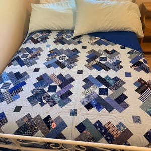 May include: A patchwork quilt featuring a log cabin block pattern in various shades of blue, navy, and white. The quilt displays a mix of floral, geometric, and nautical prints, finished with a light blue background and intricate swirling quilting.