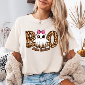 May include: A cream-colored t-shirt featuring a graphic of a ghost with a pink glitter bow and leopard print accents. The text reads BOO with leopard print letters and BASIC WITCH in brown lettering with a small brown heart icon.