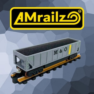May include: A gray toy hopper train car with black coal detailing, featuring the text M & O on the side and a yellow vertical stripe. The car sits on a black and orange track. Above the train is a black logo with yellow text reading AMrailz.