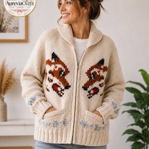 Könnte beinhalten: Eine cremefarbene Strickjacke mit Reißverschluss aus grobem Strick, ausgestattet mit einem Schalkragen und zwei Vordertaschen. Das Kleidungsstück zeigt ein wiederkehrendes Intarsienmuster aus Fuchsköpfen in Braun, Schwarz und Rot sowie hellblaue Blumenakzente an den gerippten Bündchen.