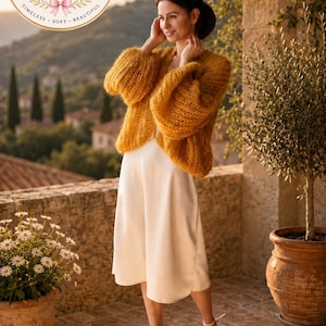 May include: A mustard yellow, open-front, long-sleeved, chunky knit cardigan with a fuzzy texture, worn over a simple, sleeveless, cream-colored midi-length slip dress. The outfit is paired with white strappy high-heeled sandals.