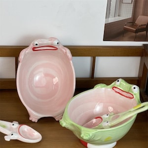 May include: Two ceramic bowls shaped like frogs, one light pink and one light green, each featuring a wide mouth with red lips and two eyes on the rim. A matching light pink spoon with a frog face design is positioned next to the bowls.