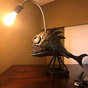 May include: A metal anglerfish lamp featuring a dark bronze-toned body with overlapping scale-like plates, sharp teeth, and a flexible silver-toned neck extending from the head to hold a glowing, warm-toned Edison-style light bulb.