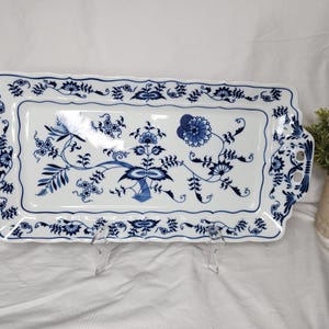 May include: A rectangular white ceramic serving tray featuring a blue onion pattern. The tray has scalloped edges, two integrated handles on the sides, and a central floral motif with intricate blue botanical designs along the border.