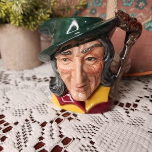 Peut inclure: Un pichet en céramique en forme de personnage portant un chapeau vert foncé. La figure a des cheveux sombres, un nez proéminent et un visage ridé. La base du pichet présente un col jaune et bordeaux, et l'anse ressemble à une canne en bois marron.