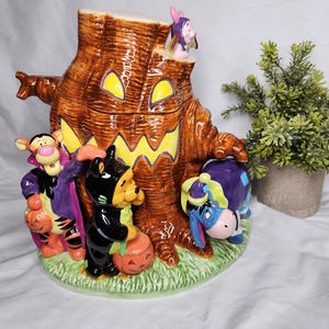 May include: A ceramic cookie jar shaped like a brown tree stump with a carved jack-o'-lantern face. Tigger, Winnie the Pooh, Eeyore, and Piglet are dressed in Halloween costumes and standing around the base of the tree stump.