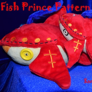 Puede incluir: Un juguete de peluche de pescado rojo y blanco con una aleta azul. El pez tiene un botón en el vientre y está cosido con hilo amarillo. El texto "Fish Prince Pattern" es visible en la parte superior de la imagen. El texto "Sample of Completed Plush" es visible en la parte inferior de la imagen.