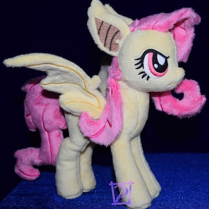 Puede incluir: Un juguete de peluche de un pony rosa y amarillo con alas de murciélago. El pony tiene una crin y una cola rosas, y sus ojos están cerrados. El pony está de pie sobre una superficie azul.