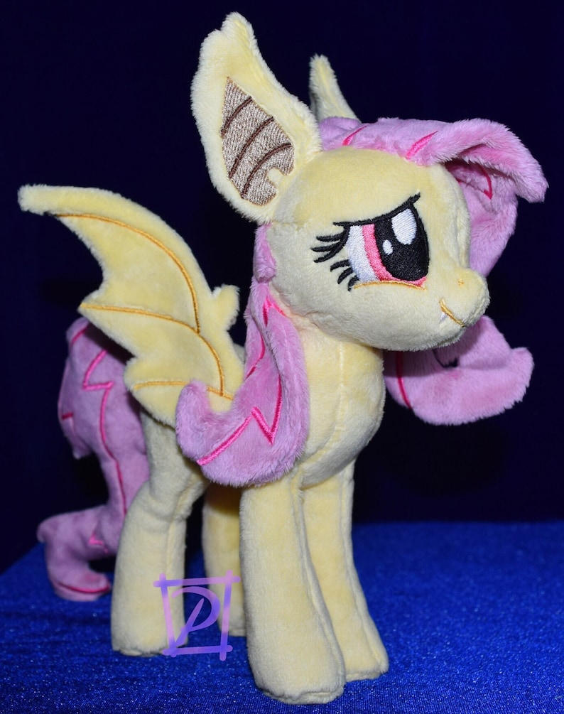 Pode incluir: Um brinquedo de pel&uacute;cia de um p&ocirc;nei amarelo e rosa com asas de morcego. O p&ocirc;nei tem uma crina e cauda rosas, e seus olhos est&atilde;o fechados. O p&ocirc;nei est&aacute; em p&eacute; sobre uma superf&iacute;cie azul.