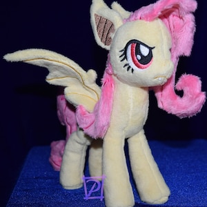 Pode incluir: Um brinquedo de pel&uacute;cia de um p&ocirc;nei rosa e amarelo com asas de morcego. O p&ocirc;nei tem uma crina e uma cauda rosas, e seus olhos s&atilde;o vermelhos. O p&ocirc;nei est&aacute; em p&eacute; sobre uma superf&iacute;cie azul.