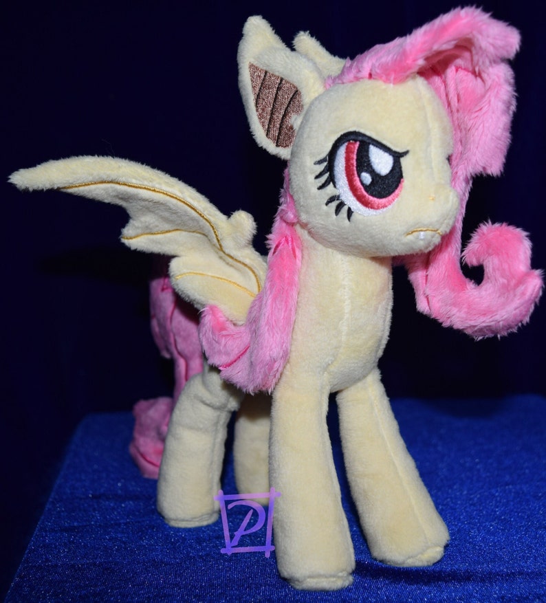 Pode incluir: Um brinquedo de pel&uacute;cia de um p&ocirc;nei rosa e amarelo com asas de morcego. O p&ocirc;nei tem uma crina e uma cauda rosas, e seus olhos s&atilde;o vermelhos. O p&ocirc;nei est&aacute; em p&eacute; sobre uma superf&iacute;cie azul.