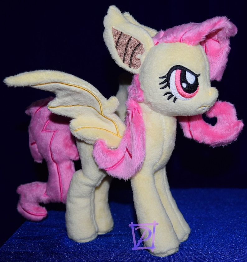 Pode incluir: Um brinquedo de pel&uacute;cia de um p&ocirc;nei rosa e amarelo com asas de morcego. O p&ocirc;nei tem uma crina e uma cauda rosas, e seus olhos est&atilde;o fechados. O p&ocirc;nei est&aacute; em p&eacute; sobre uma superf&iacute;cie azul.