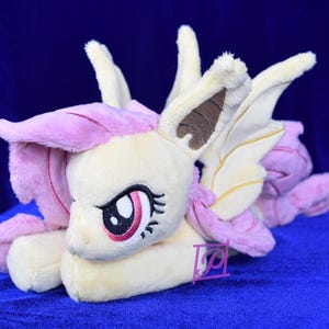 Pode incluir: Um brinquedo de pel&uacute;cia de um poney rosa e amarelo com orelhas e asas marrons. O poney tem uma crina e uma cauda rosas e est&aacute; deitado de lado com a cabe&ccedil;a apoiada nas patas.