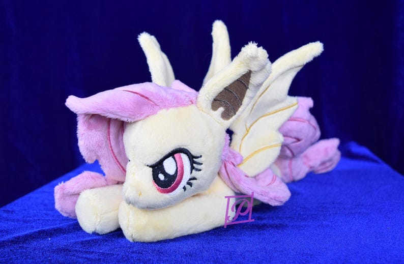Pode incluir: Um brinquedo de pel&uacute;cia de um poney rosa e amarelo com orelhas e asas marrons. O poney tem uma crina e uma cauda rosas e est&aacute; deitado de lado com a cabe&ccedil;a apoiada nas patas.