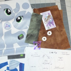 Puede incluir: Un patrón de costura para un juguete de peluche, con instrucciones para hacer un personaje de Little Pip. El patrón incluye piezas para la cola, la melena, el flequillo y el cuerpo. El patrón está impreso en papel blanco con tinta negra.