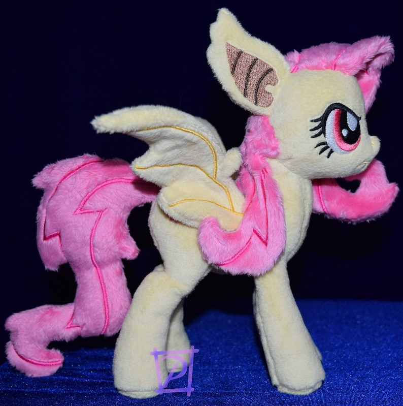 Pode incluir: Um brinquedo de pel&uacute;cia de um p&ocirc;nei amarelo e rosa com asas de morcego. O p&ocirc;nei tem uma crina e uma cauda rosas, e seus olhos s&atilde;o vermelhos. O p&ocirc;nei est&aacute; em p&eacute; sobre uma superf&iacute;cie azul.