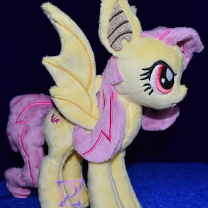 Pode incluir: Um brinquedo de pel&uacute;cia de um p&ocirc;nei amarelo e rosa com asas de morcego. O p&ocirc;nei tem uma crina e uma cauda rosas, e suas asas s&atilde;o de um amarelo claro com detalhes marrons.