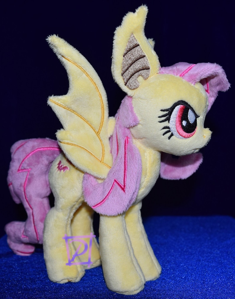 Pode incluir: Um brinquedo de pel&uacute;cia de um p&ocirc;nei amarelo e rosa com asas de morcego. O p&ocirc;nei tem uma crina e uma cauda rosas, e suas asas s&atilde;o de um amarelo claro com detalhes marrons.