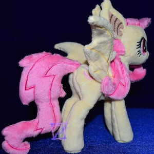 Pode incluir: Um brinquedo de pel&uacute;cia de um p&ocirc;nei amarelo e rosa com asas e uma cauda rosa. O p&ocirc;nei tem uma crina rosa e uma cauda rosa com um design de raio. O p&ocirc;nei tem um nariz rosa e olhos vermelhos.