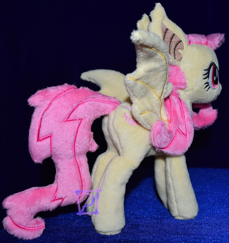 Pode incluir: Um brinquedo de pel&uacute;cia de um p&ocirc;nei amarelo e rosa com asas e uma cauda rosa. O p&ocirc;nei tem uma crina rosa e uma cauda rosa com um design de raio. O p&ocirc;nei tem um nariz rosa e olhos vermelhos.