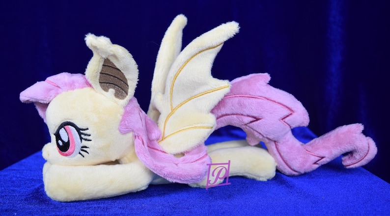 Pode incluir: Um brinquedo de pel&uacute;cia de um p&ocirc;nei alado com um corpo marrom claro, uma crina e uma cauda rosa e asas douradas. O p&ocirc;nei est&aacute; deitado de lado com a cabe&ccedil;a apoiada nas patas dianteiras.