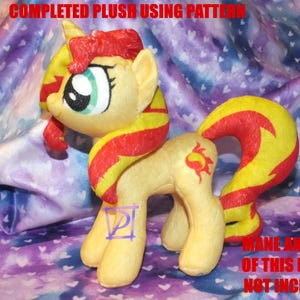 May include: A completed plush toy of a yellow pony with a red and yellow mane and tail. The pony has a green eye and a sun symbol on its flank. The plush toy is sitting on a purple and white fabric with a heart pattern.