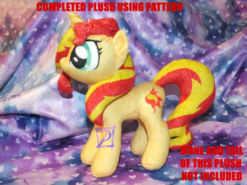 May include: A completed plush toy of a yellow pony with a red and yellow mane and tail. The pony has a green eye and a sun symbol on its flank. The plush toy is sitting on a purple and white fabric with a heart pattern.
