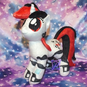 May include: A white and black striped plush pony with a red and black mane and tail. The pony has a red ribbon around its neck and a black and white symbol on its flank.