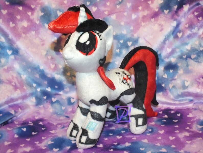 May include: A white and black striped plush pony with a red and black mane and tail. The pony has a red ribbon around its neck and a black and white symbol on its flank.