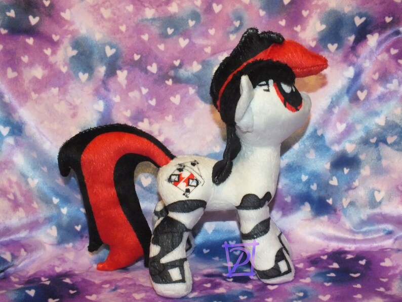 May include: A black and red plush toy pony with a white body and black and white markings. The pony has a red and black mane and tail, and a red bandana on its head. The pony has a black and white design on its flank that includes playing cards.