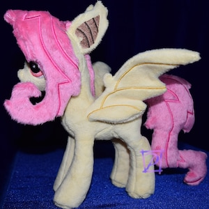 Pode incluir: Um brinquedo de pel&uacute;cia de um p&ocirc;nei alado com cabelo rosa e corpo amarelo. O p&ocirc;nei tem uma cauda encaracolada e uma &uacute;nica asa nas costas.