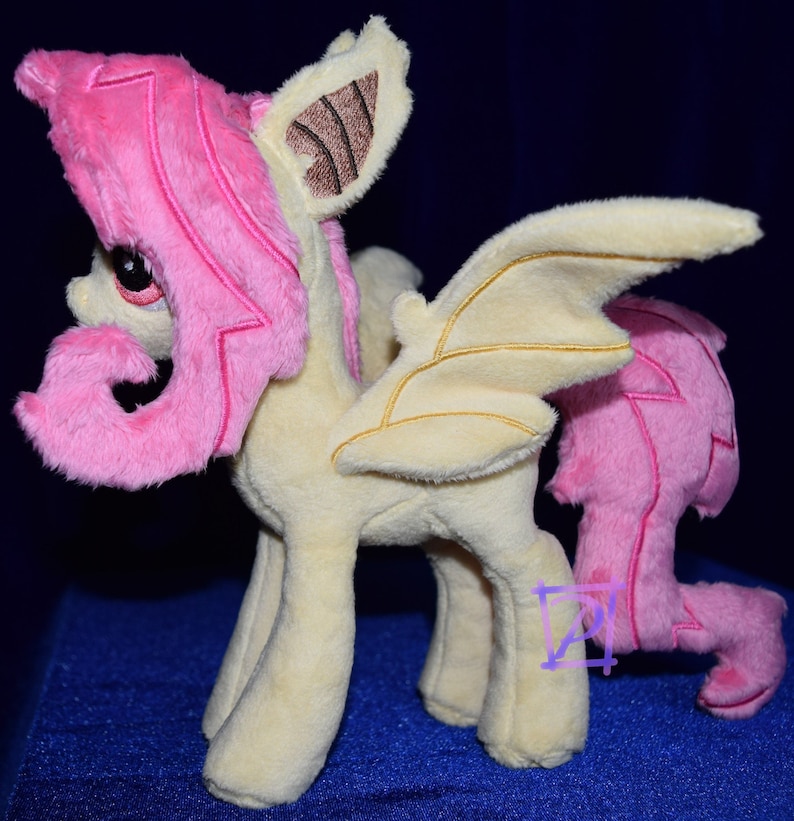 Pode incluir: Um brinquedo de pel&uacute;cia de um p&ocirc;nei alado com cabelo rosa e corpo amarelo. O p&ocirc;nei tem uma cauda encaracolada e uma &uacute;nica asa nas costas.
