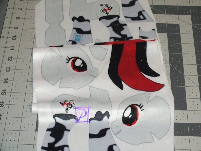 May include: White fabric with a pattern of a cartoon pony with a red eye and black mane. The fabric is cut into different shapes for sewing a plush toy.