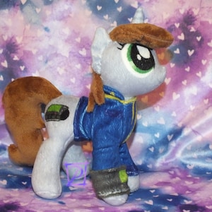 Könnte beinhalten: Ein Plüsch-Pony mit hellgrauem Fell, brauner Mähne und Schweif sowie einem Horn. Das Pony trägt eine blaue Jacke mit gelbem Saum, eine braune Mütze und eine dunkelgraue Manschette an einem Bein. Es hat große grüne Augen und ein grünes Detail.