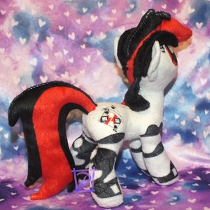 May include: A plush toy pony with a black and red mane and tail. The pony is wearing a black and white striped outfit with a red playing card on its back. The pony is standing on a purple and blue background with white hearts.