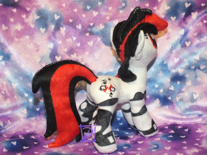 May include: A plush toy pony with a black and red mane and tail. The pony is wearing a black and white striped outfit with a red playing card on its back. The pony is standing on a purple and blue background with white hearts.