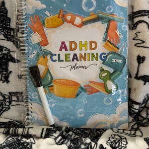 May include: A spiral-bound ADHD Cleaning planner with a light blue cover featuring illustrations of cleaning supplies like orange gloves, sponges, and spray bottles. The text reads ADHD CLEANING planner. A black dry-erase marker is attached to the front.