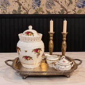 May include: A white ceramic biscuit jar with a lid, a small creamer, and a sugar bowl, all decorated with red and yellow rose patterns and gold trim. These items sit on an ornate silver-toned metal serving tray with two tall candlesticks.