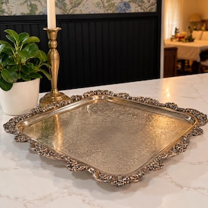 May include: A square silver-toned serving tray featuring an intricate embossed floral pattern on the central surface and a decorative scalloped edge with detailed scrollwork along the entire perimeter of the metal frame.