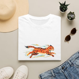 Op de afbeelding: Een wit T-shirt met korte mouwen voorzien van een grafische illustratie van een oranje en zwart gestreepte kat die op hoge snelheid rent, afgebeeld met gestileerde bewegingslijnen in tinten blauwgroen en geel rondom het katachtige figuur.
