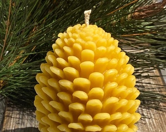 May include: A yellow beeswax candle shaped like a pine cone. The candle has a white wick and is sitting on a wooden surface.