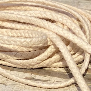 May include: A close-up of a spool of natural white braided cotton cord. The cord is soft and pliable, ideal for crafting and macrame projects.