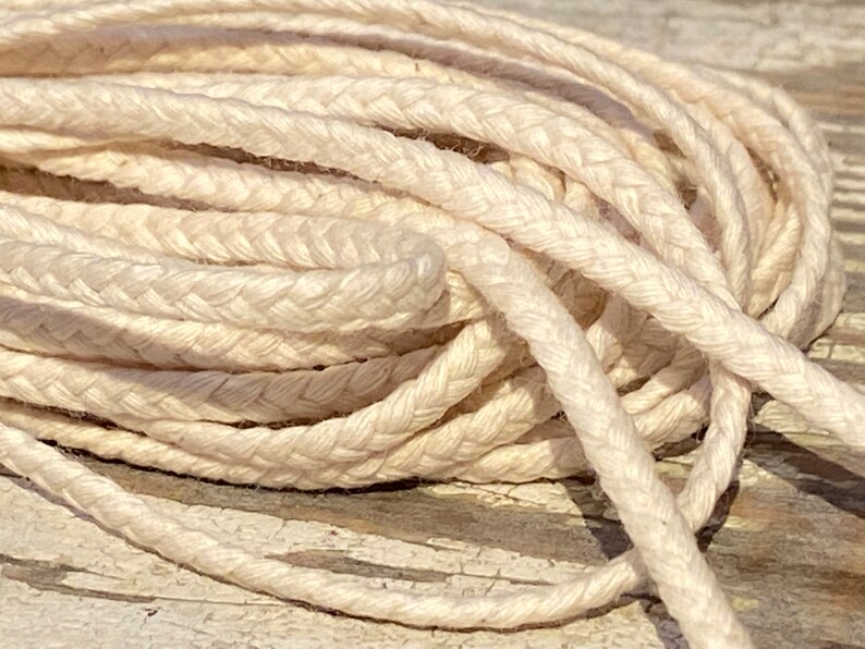 May include: A close-up of a spool of natural white braided cotton cord. The cord is soft and pliable, ideal for crafting and macrame projects.