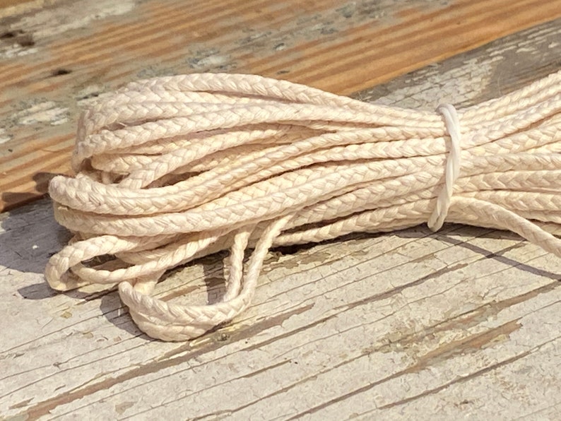 May include: A close-up of a white braided cotton cord, tied with a rubber band. The cord is on a wooden surface.
