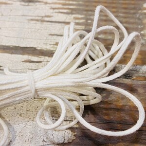 May include: A close-up of a white cotton cord, twisted and coiled on a wooden surface. The cord is soft and pliable, perfect for crafting or macrame projects.
