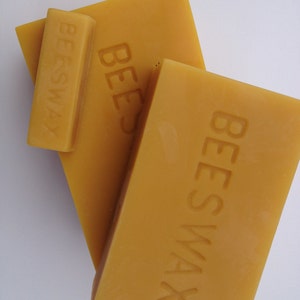 May include: Two blocks of yellow beeswax, one on top of the other. The top block has the word "BEESWAX" embossed on it. The bottom block has the word "BEES" embossed on it. A smaller block of beeswax with the word "BEESWAX" embossed on it is sitting on top of the bottom block.
